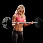 Fit blonde woman in pink top and black leggings performing bicep curls with a barbell