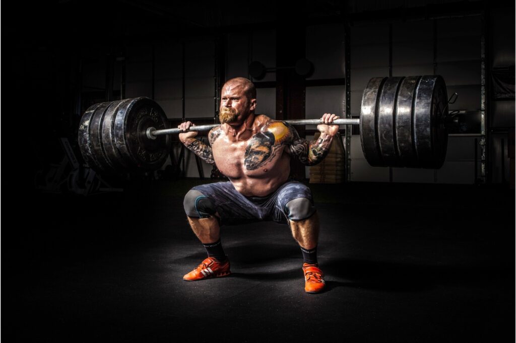 Muscular bald man with beard and chest tattoo squatting with heavy weights