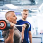 Senior man performing bicep curls with a barbell under the supervision of a personal trainer